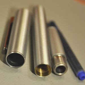 Może przedstawiać: Rozłożony srebrny długopis, ukazujący jego wewnętrzne elementy. Widoczne są skuwka, korpus, wkład atramentowy i końc&oacute;wka. Wnętrze korpusu jest złote. Długopis leży na neutralnej powierzchni.