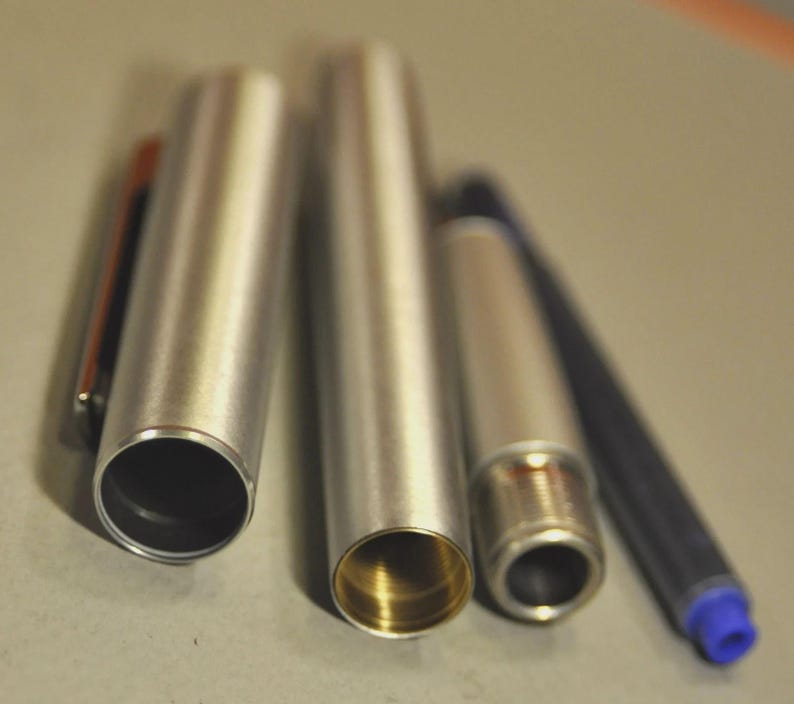 Może przedstawiać: Rozłożony srebrny długopis, ukazujący jego wewnętrzne elementy. Widoczne są skuwka, korpus, wkład atramentowy i końc&oacute;wka. Wnętrze korpusu jest złote. Długopis leży na neutralnej powierzchni.