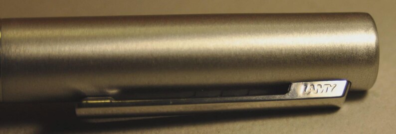 May include: Close-up of a silver-colored pen with a clip. The pen's body has a brushed metal finish. The clip is rectangular with the brand name "LAMY" engraved on it.