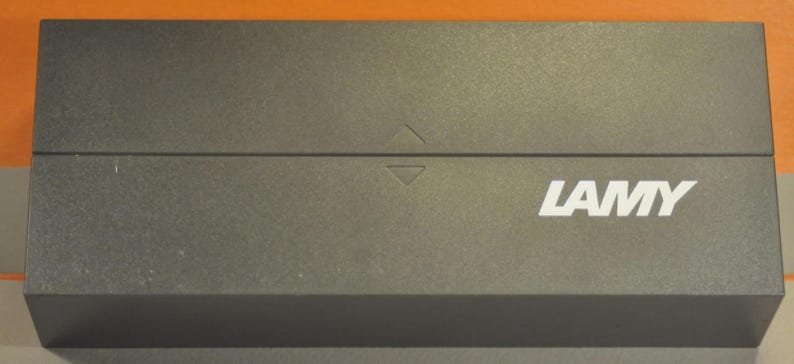 May include: A rectangular, dark gray box with the white word "LAMY" printed on the front. The box has a textured surface and a hinged lid. A small arrow is visible on the top of the box.