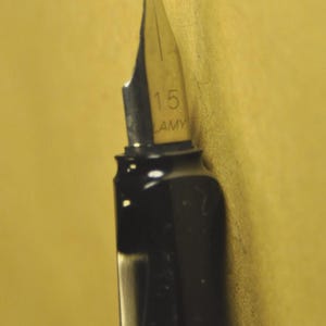 K&ouml;nnte beinhalten: Nahaufnahme einer schwarzen F&uuml;llfederhalterfeder mit silberfarbener Spitze. Die Feder ist mit "1.5" und "LAMY" gekennzeichnet. Der Stift befindet sich vor einem gelben Hintergrund.
