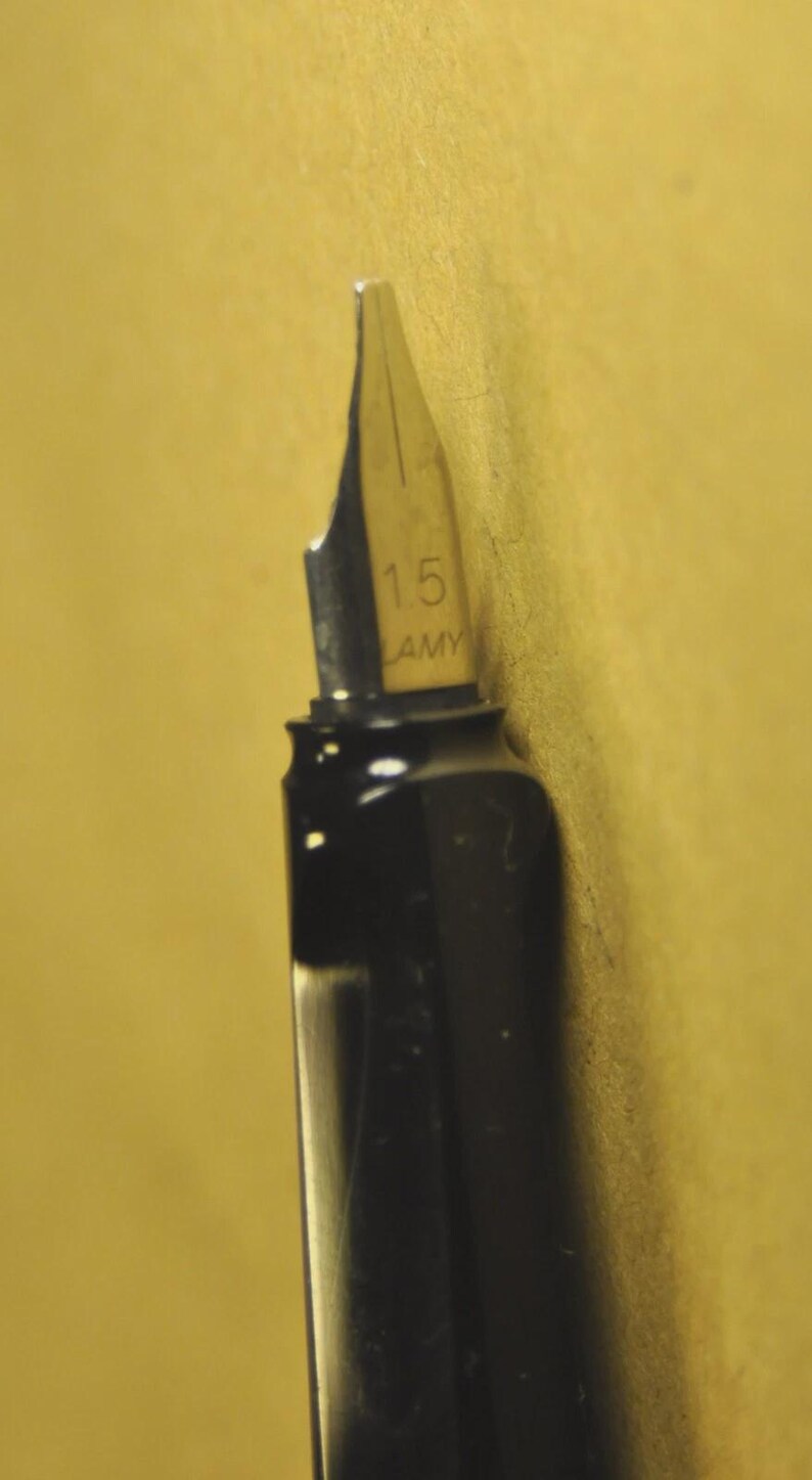 K&ouml;nnte beinhalten: Nahaufnahme einer schwarzen F&uuml;llfederhalterfeder mit silberfarbener Spitze. Die Feder ist mit "1.5" und "LAMY" gekennzeichnet. Der Stift befindet sich vor einem gelben Hintergrund.