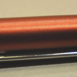 May include: A metallic copper-colored fountain pen featuring a smooth cylindrical barrel and a polished silver-tone metal clip. The clip is engraved with the brand name Lamy in a clear, legible font, positioned against the matte finish of the pen body.
