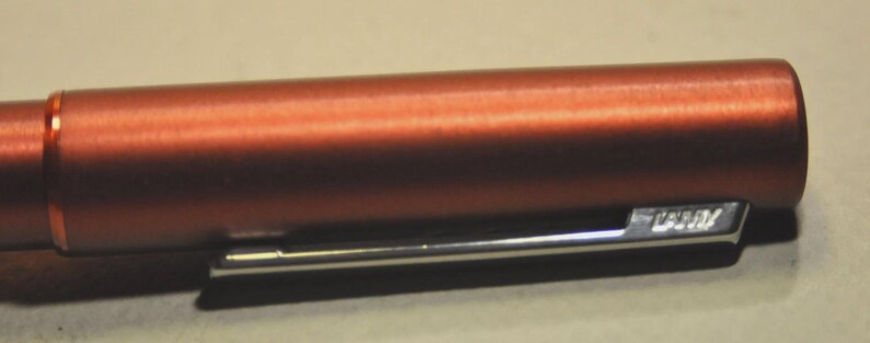 May include: A metallic copper-colored fountain pen featuring a smooth cylindrical barrel and a polished silver-tone metal clip. The clip is engraved with the brand name Lamy in a clear, legible font, positioned against the matte finish of the pen body.