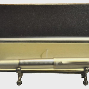 Op de afbeelding: Een zilverkleurige pen ligt in een open zwarte en crèmekleurige pennendoos. De pen heeft een geribbelde grip en een gebogen clip. De binnenkant van de doos is crèmekleurig. De pen wordt op een decoratieve standaard gepresenteerd.