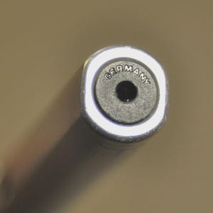 K&ouml;nnte beinhalten: Ein zylindrisches Metallobjekt mit einer runden schwarzen Endkappe, die in der Mitte ein Loch aufweist. Das Wort GERMANY ist bogenf&ouml;rmig am oberen Rand des schwarzen Einsatzes eingepr&auml;gt, der von einem glatten, reflektierenden Silberrand umgeben ist.