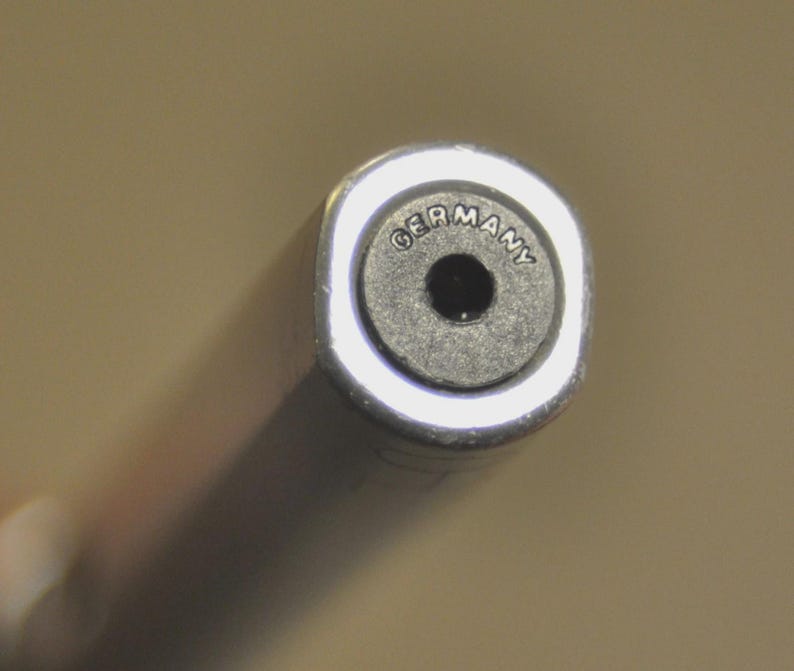 K&ouml;nnte beinhalten: Ein zylindrisches Metallobjekt mit einer runden schwarzen Endkappe, die in der Mitte ein Loch aufweist. Das Wort GERMANY ist bogenf&ouml;rmig am oberen Rand des schwarzen Einsatzes eingepr&auml;gt, der von einem glatten, reflektierenden Silberrand umgeben ist.
