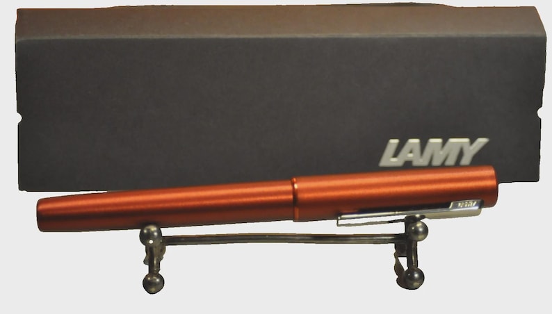 May include: A metallic orange fountain pen with a silver clip rests on a small metal stand in front of a dark gray rectangular box. The word LAMY is printed in white on the right side of the box, which serves as a storage case for the pen.