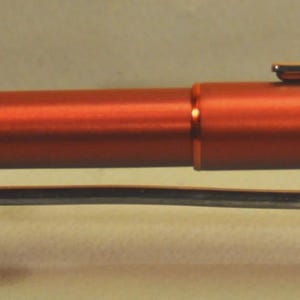 May include: A metallic burnt orange fountain pen with a smooth, cylindrical body and a matching cap. The pen features a slender, polished silver-tone metal clip attached to the cap and rests horizontally on a small, silver-colored metal stand.