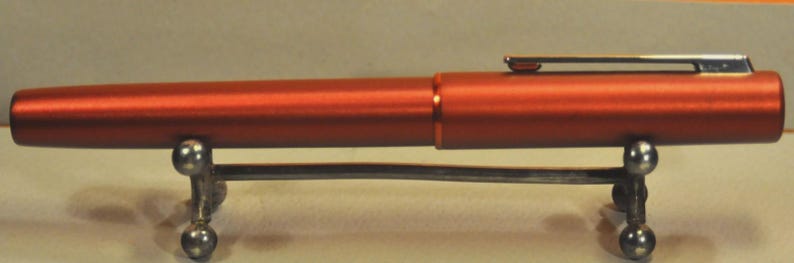 May include: A metallic burnt orange fountain pen with a smooth, cylindrical body and a matching cap. The pen features a slender, polished silver-tone metal clip attached to the cap and rests horizontally on a small, silver-colored metal stand.