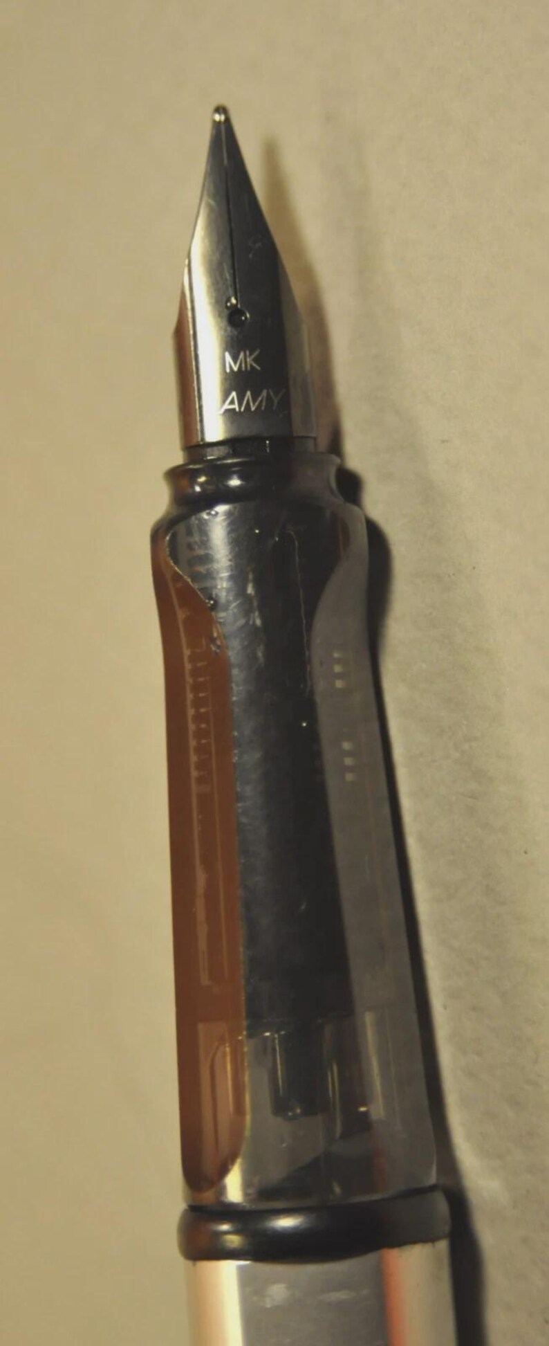 K&ouml;nnte beinhalten: Ein F&uuml;llfederhalter mit einer silberfarbenen Metallfeder, die die Gravur MK AMY tr&auml;gt. Der Stift hat ein transparentes, dunkelbraunes Griffst&uuml;ck, das innere Komponenten erkennen l&auml;sst, und einen schwarzen Schaft mit glattem, zylindrischem Design.
