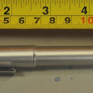 May include: A silver pen with a clip, approximately 5.5 inches long, is positioned next to a yellow tape measure. The pen has a cylindrical body and a cap. The tape measure shows measurements in inches and feet.