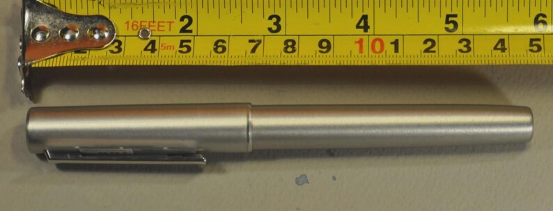 May include: A silver pen with a clip, approximately 5.5 inches long, is positioned next to a yellow tape measure. The pen has a cylindrical body and a cap. The tape measure shows measurements in inches and feet.
