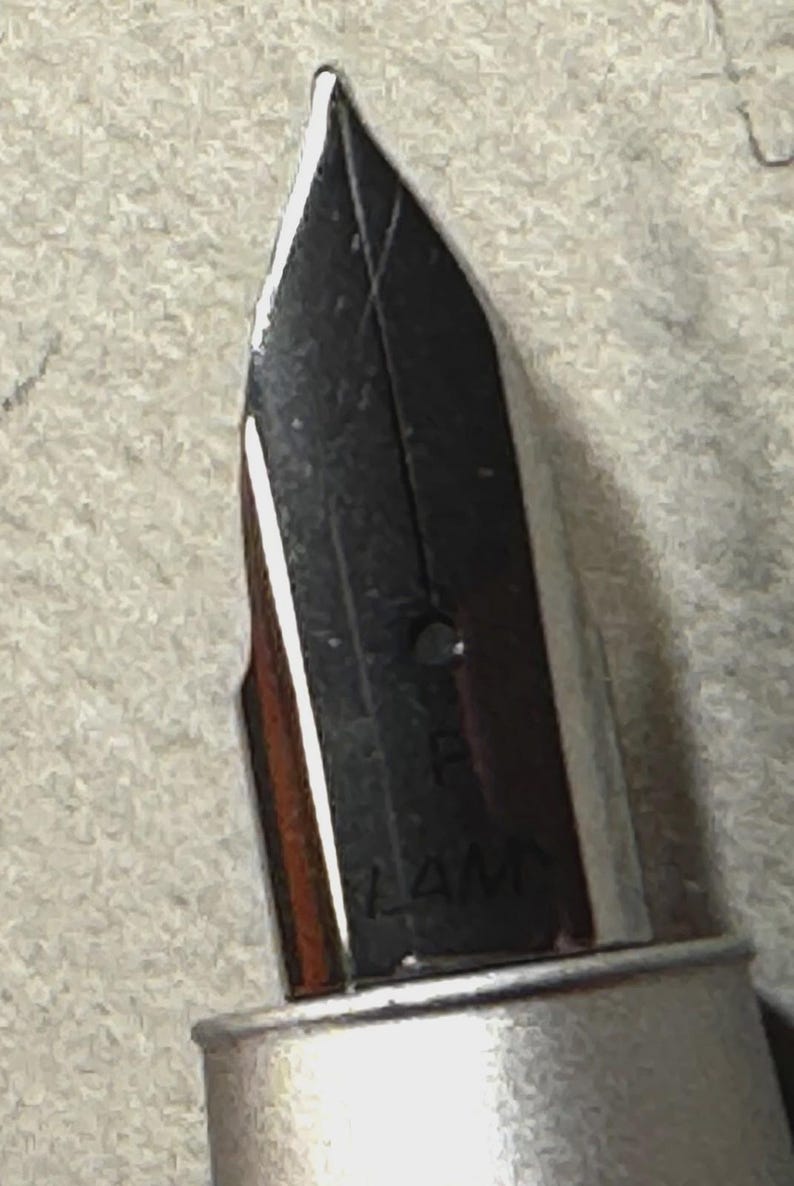 Może przedstawiać: Zbliżenie srebrnej stal&oacute;wki pi&oacute;ra wiecznego. Stal&oacute;wka jest czarna z cienką końc&oacute;wką i widoczną nazwą marki "LAMY". Pi&oacute;ro ma metaliczny srebrny korpus.