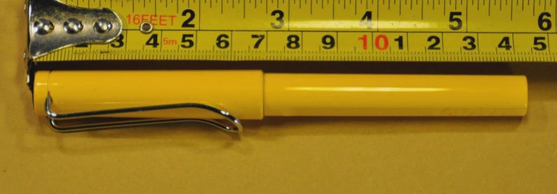 Puede incluir: Un bol&iacute;grafo amarillo con un clip plateado, de aproximadamente 14 cm de largo, est&aacute; colocado junto a una cinta m&eacute;trica amarilla y negra. El bol&iacute;grafo es cil&iacute;ndrico con una tapa. La cinta m&eacute;trica muestra medidas en pies y pulgadas.
