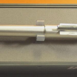 May include: A silver-colored Lamy pen resting inside its gray box. The pen is cylindrical with a clip and a label attached. The box has the brand name "LAMY" printed on it.