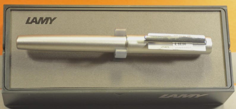 May include: A silver-colored Lamy pen resting inside its gray box. The pen is cylindrical with a clip and a label attached. The box has the brand name "LAMY" printed on it.