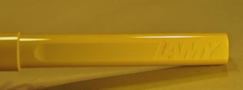 Puede incluir: Primer plano de un barril de bol&iacute;grafo amarillo brillante con el nombre de la marca "LAMY" grabado en relieve en el lateral. El bol&iacute;grafo tiene un acabado liso y brillante y una forma cil&iacute;ndrica. El fondo es amarillo liso y apagado.