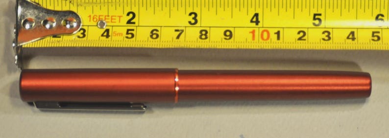 May include: A metallic rust-colored pen with a silver clip, positioned below a yellow measuring tape that shows the pen is approximately 5.5 inches in length. The pen features a smooth, cylindrical body with a thin orange ring at the center.