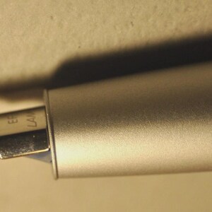 May include: Close-up of a silver fountain pen with a metallic finish. The nib is visible, with the brand name "EF LAMY" engraved on it. The pen is positioned at an angle, casting a shadow on the background.