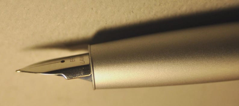 May include: Close-up of a silver fountain pen with a metallic finish. The nib is visible, with the brand name "EF LAMY" engraved on it. The pen is positioned at an angle, casting a shadow on the background.