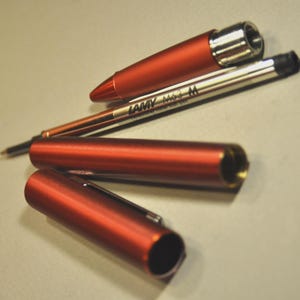 May include: A disassembled metallic orange pen consisting of a cap, a cylindrical body, and a front section. A silver LAMY M63 M rollerball pen refill is positioned diagonally across the components, clearly displaying the brand and model text.
