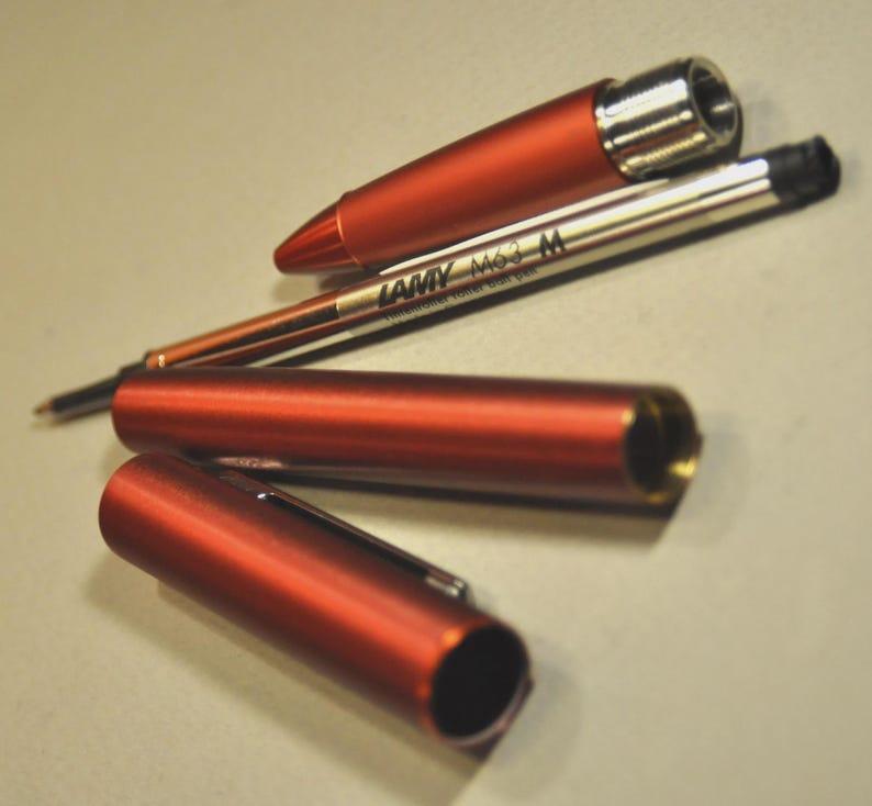 May include: A disassembled metallic orange pen consisting of a cap, a cylindrical body, and a front section. A silver LAMY M63 M rollerball pen refill is positioned diagonally across the components, clearly displaying the brand and model text.