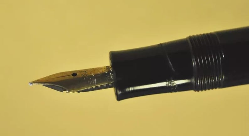 Puede incluir: Una pluma estilogr&aacute;fica negra con un plum&iacute;n met&aacute;lico de tono dorado que presenta un intrincado dise&ntilde;o grabado. El plum&iacute;n est&aacute; unido a una secci&oacute;n de agarre negra, lisa y brillante, que se conecta a un cuerpo roscado de dise&ntilde;o cl&aacute;sico.