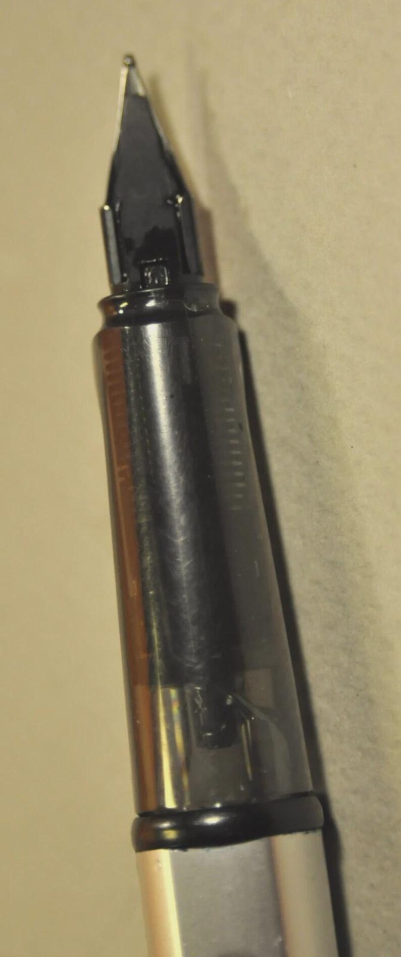 K&ouml;nnte beinhalten: Ein F&uuml;llfederhalter mit einem unteren Schaft aus silberfarbenem Metall und einem Griffst&uuml;ck aus durchscheinendem Dunkelgrau. Die Feder ist dunkel gef&auml;rbt und spitz zulaufend. Der interne Tintenleiter ist durch das halbtransparente Material sichtbar.