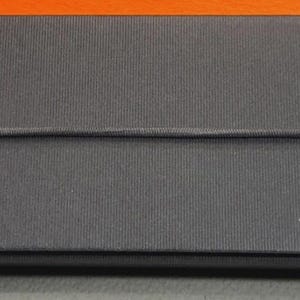 Może przedstawiać: Prostokątne, ciemnoszare pudełko z białym napisem "LAMY" z przodu. Pudełko ma teksturowaną powierzchnię i cienką, ciemną gumkę owiniętą wok&oacute;ł niego. Tło jest pomarańczowe i szare.