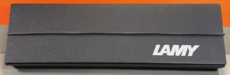 Może przedstawiać: Prostokątne, ciemnoszare pudełko z białym napisem "LAMY" z przodu. Pudełko ma teksturowaną powierzchnię i cienką, ciemną gumkę owiniętą wok&oacute;ł niego. Tło jest pomarańczowe i szare.