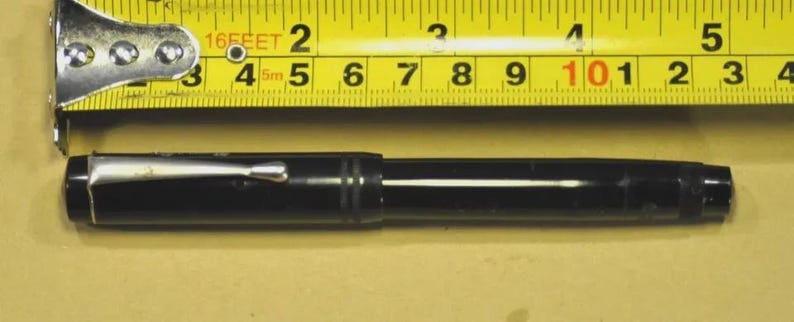 Puede incluir: Una pluma estilogr&aacute;fica negra con un clip de metal plateado colocada debajo de una cinta m&eacute;trica amarilla. La pluma mide aproximadamente 13,3 cm de largo, con un cuerpo cil&iacute;ndrico liso y detalles de anillos horizontales cerca de la tapa.
