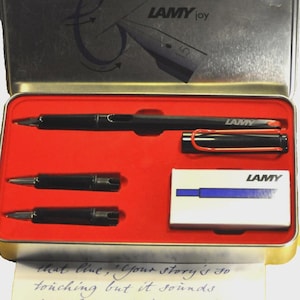 K&ouml;nnte beinhalten: Ein schwarzes Lamy Joy F&uuml;llfederhalter-Set in einer silbernen Metallbox mit rotem Innenraum. Das Set enth&auml;lt den Stift, zwei Federn und eine kleine Schachtel. Das Etui tr&auml;gt das Lamy-Logo und den Text "LAMY joy".