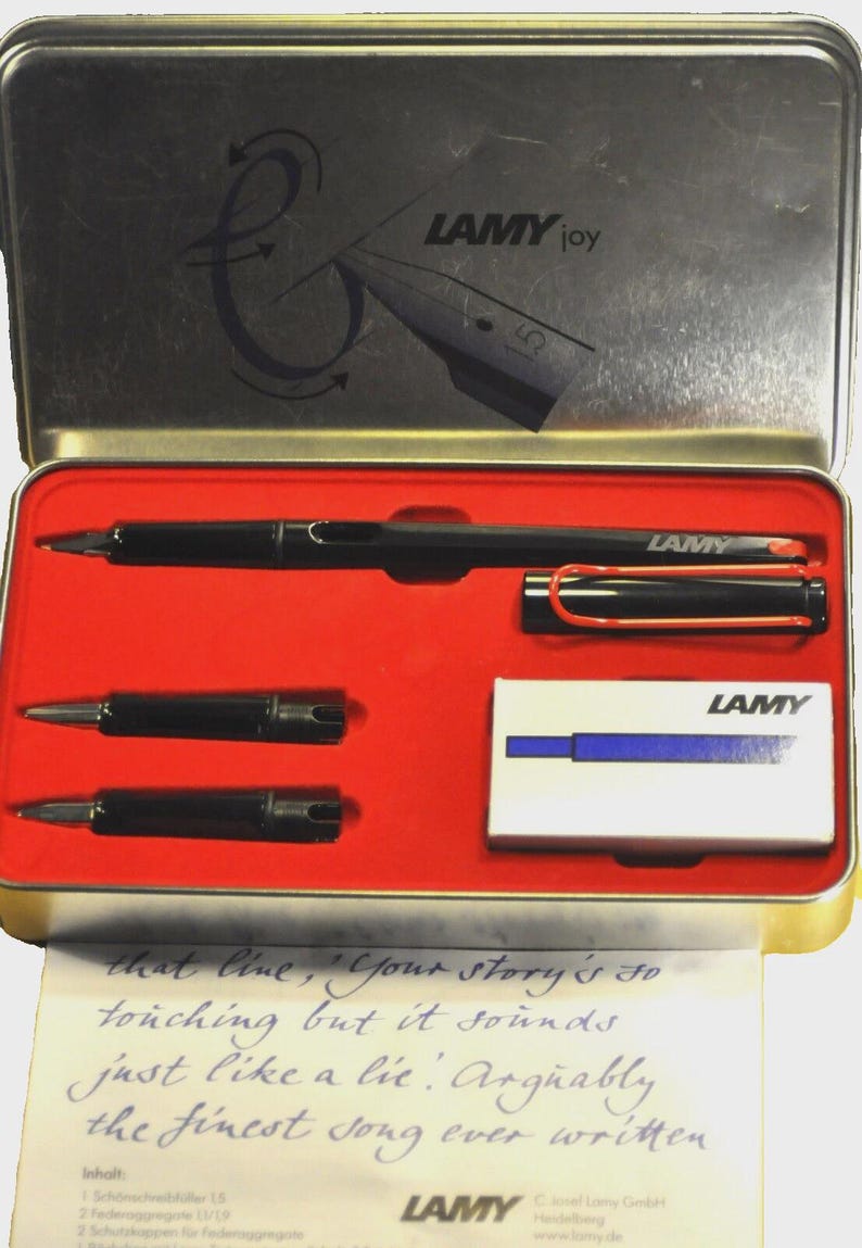 K&ouml;nnte beinhalten: Ein schwarzes Lamy Joy F&uuml;llfederhalter-Set in einer silbernen Metallbox mit rotem Innenraum. Das Set enth&auml;lt den Stift, zwei Federn und eine kleine Schachtel. Das Etui tr&auml;gt das Lamy-Logo und den Text "LAMY joy".