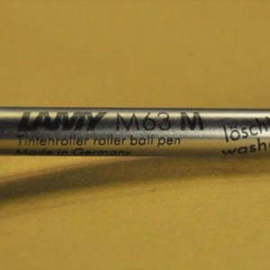 Puede incluir: Un cartucho de tinta plateado Lamy M63 M para bol&iacute;grafo roller. El cartucho tiene texto negro que dice "Lamy M63 M Tintensroller ball pen" y "l&ouml;schbar washable". La punta es azul.