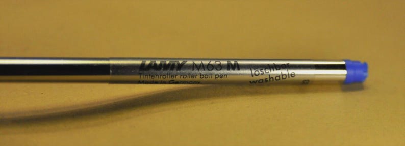 Puede incluir: Un cartucho de tinta plateado Lamy M63 M para bol&iacute;grafo roller. El cartucho tiene texto negro que dice "Lamy M63 M Tintensroller ball pen" y "l&ouml;schbar washable". La punta es azul.