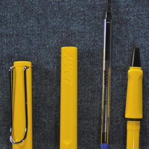 Puede incluir: Un bol&iacute;grafo LAMY amarillo desmontado con un clip plateado, un capuch&oacute;n amarillo con el logotipo de LAMY, un cartucho de tinta negro y plateado y un cuerpo de bol&iacute;grafo amarillo con punta negra. El bol&iacute;grafo est&aacute; sobre un fondo vaquero.