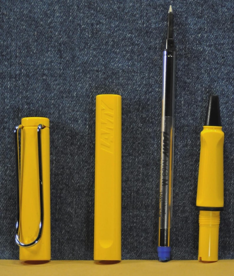 Puede incluir: Un bol&iacute;grafo LAMY amarillo desmontado con un clip plateado, un capuch&oacute;n amarillo con el logotipo de LAMY, un cartucho de tinta negro y plateado y un cuerpo de bol&iacute;grafo amarillo con punta negra. El bol&iacute;grafo est&aacute; sobre un fondo vaquero.