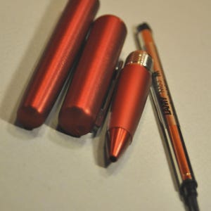 May include: A disassembled metallic red pen consisting of a cylindrical cap, a barrel, a tapered front section, and a Lamy M63 M ink refill. The components are arranged side-by-side, showcasing the matte finish of the red metal casing and the ink cartridge.