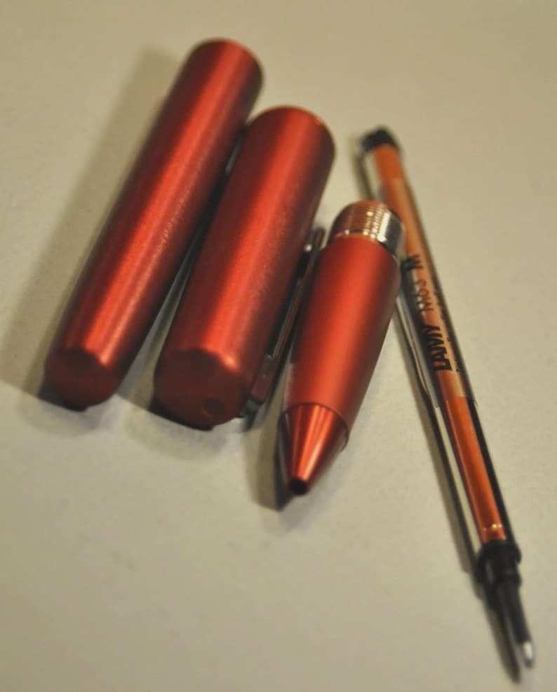 May include: A disassembled metallic red pen consisting of a cylindrical cap, a barrel, a tapered front section, and a Lamy M63 M ink refill. The components are arranged side-by-side, showcasing the matte finish of the red metal casing and the ink cartridge.