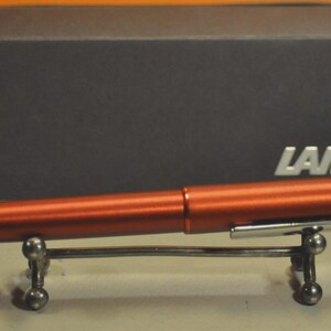 May include: A metallic copper-colored fountain pen with a silver-toned clip featuring the brand name LAMY. The pen rests on a small silver metal stand. Behind the pen sits a dark gray rectangular box with the white text LAMY printed on the front.