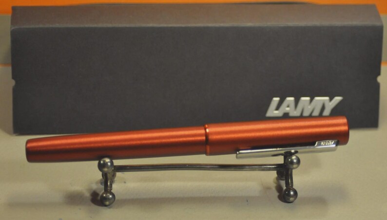 May include: A metallic copper-colored fountain pen with a silver-toned clip featuring the brand name LAMY. The pen rests on a small silver metal stand. Behind the pen sits a dark gray rectangular box with the white text LAMY printed on the front.