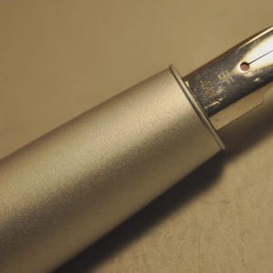 May include: Close-up of a silver fountain pen with a metallic finish. The pen's nib is visible, showcasing its design and the fine point. The pen is set against a neutral background, highlighting its sleek, modern aesthetic. A writing instrument.