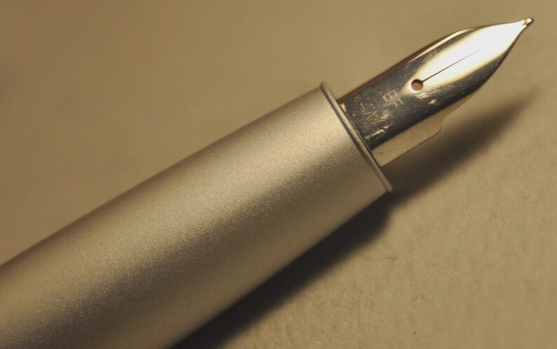 May include: Close-up of a silver fountain pen with a metallic finish. The pen's nib is visible, showcasing its design and the fine point. The pen is set against a neutral background, highlighting its sleek, modern aesthetic. A writing instrument.