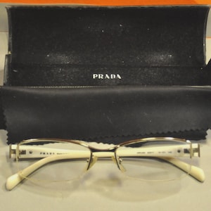 May include: A pair of Prada eyeglasses with a gold-colored frame and off-white arms. The glasses are displayed with a black case and a black cleaning cloth. The Prada logo is visible on the case and the glasses.
