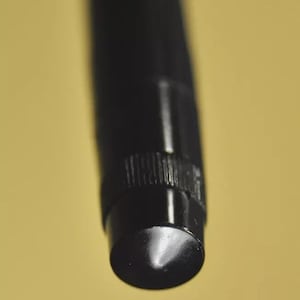 Puede incluir: Un instrumento de escritura cil&iacute;ndrico de color negro con un acabado mate liso y una secci&oacute;n de agarre texturizada cerca de la punta. La punta es redondeada y se estrecha, mostrando un brillo lustroso donde la luz se refleja en la superficie.