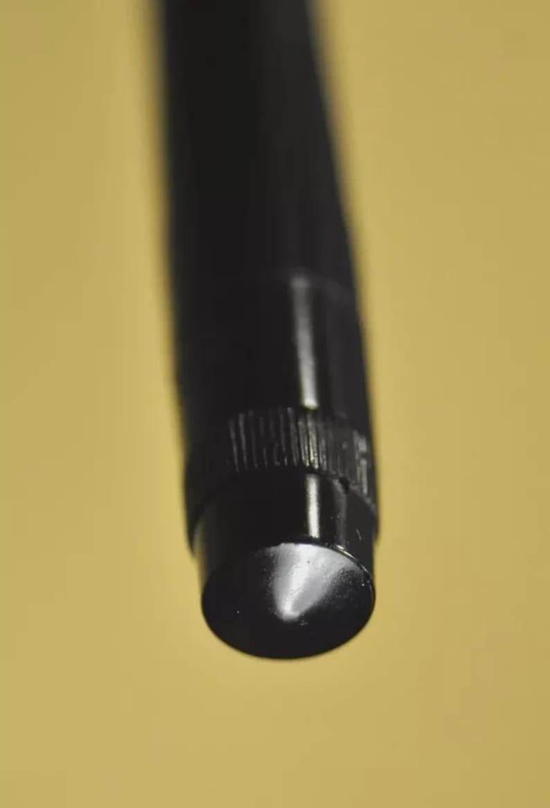 Puede incluir: Un instrumento de escritura cil&iacute;ndrico de color negro con un acabado mate liso y una secci&oacute;n de agarre texturizada cerca de la punta. La punta es redondeada y se estrecha, mostrando un brillo lustroso donde la luz se refleja en la superficie.