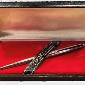 May include: A silver pen and a black pen sleeve with gold lettering are displayed inside a black box with a red velvet interior. The pen sleeve reads "MADISON by CARAN d'ACHE". The box is open, revealing the pen and sleeve.