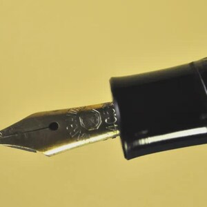Puede incluir: Un plum&iacute;n de pluma estilogr&aacute;fica hecho de metal dorado con un dise&ntilde;o grabado y una ranura central, unido a una secci&oacute;n de agarre de pl&aacute;stico negro. El plum&iacute;n presenta un peque&ntilde;o orificio de ventilaci&oacute;n circular y marcas grabadas.