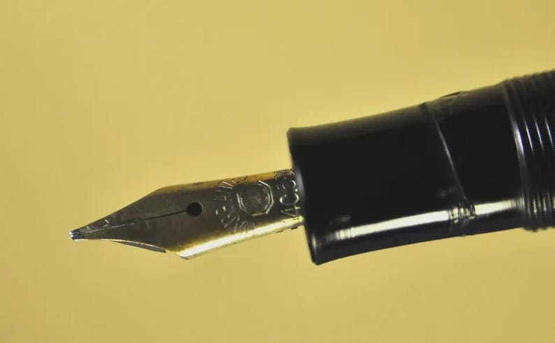Puede incluir: Un plum&iacute;n de pluma estilogr&aacute;fica hecho de metal dorado con un dise&ntilde;o grabado y una ranura central, unido a una secci&oacute;n de agarre de pl&aacute;stico negro. El plum&iacute;n presenta un peque&ntilde;o orificio de ventilaci&oacute;n circular y marcas grabadas.